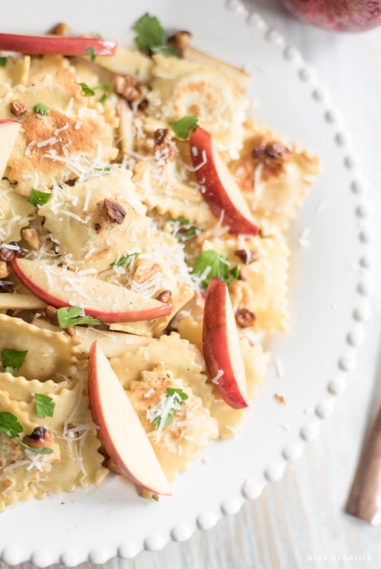 Closeup of a white plate featuring apples, ravioli, parmesan cheese, and parsley.