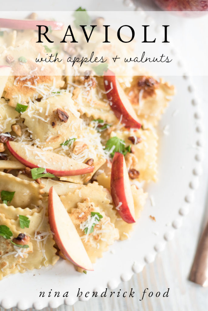 Closeup of a white plate featuring apples, ravioli, parmesan cheese, and parsley.